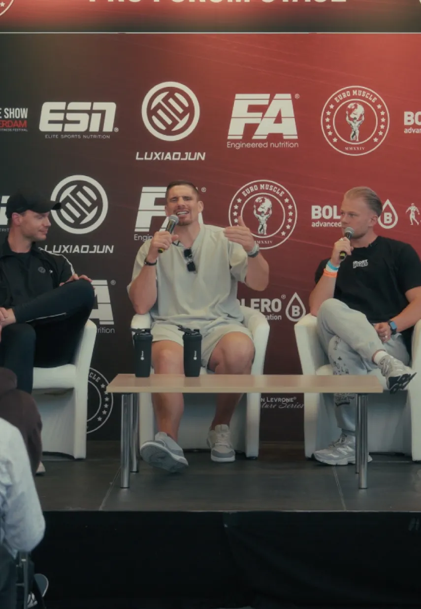 Drie mannen zitten op een podium tijdens een fitness paneldiscussie met microfoons in de hand, voor een backdrop met sportmerken.