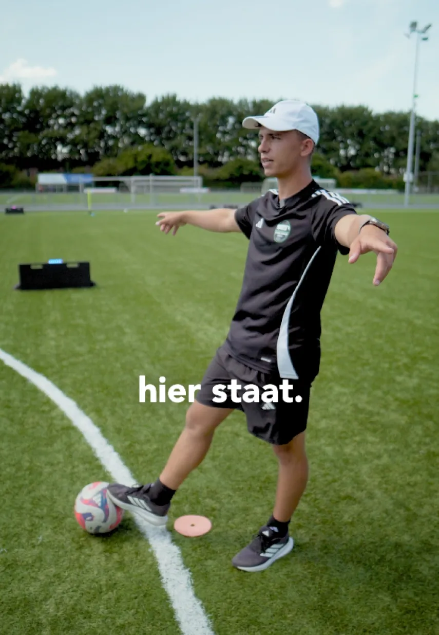 Voetbaltrainer op kunstgrasveld wijst naar een plek op de grond met tekst 'hier staat.' in beeld.