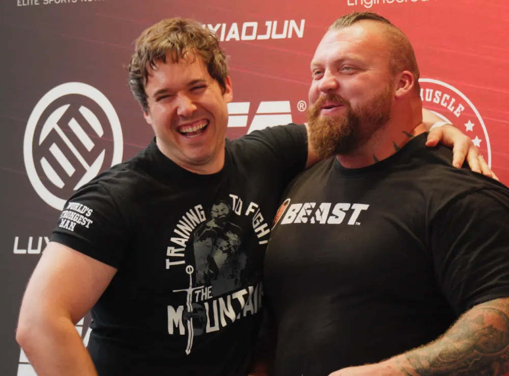 Twee gespierde mannen lachen en omhelzen elkaar voor een backdrop met sportmerken, één draagt een 'Beast'-shirt.