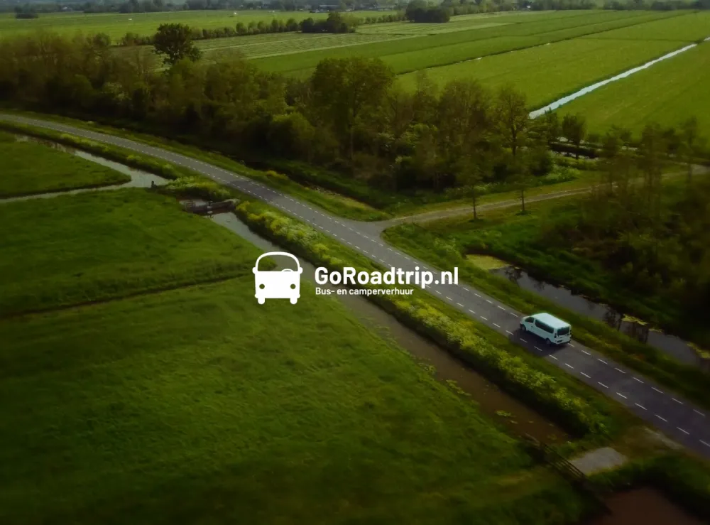 Dronebeeld van een witte bus die over een kronkelende weg door een groen landschap rijdt, met GoRoadtrip.nl logo.