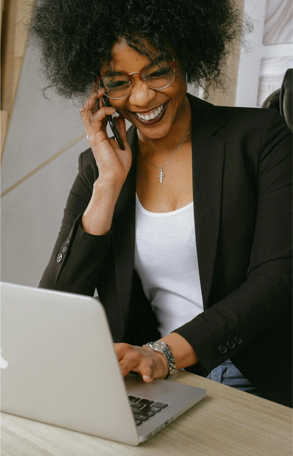 A smiling woman wearing glasses and a black blazer is talking on the phone while working on a laptop at a desk.