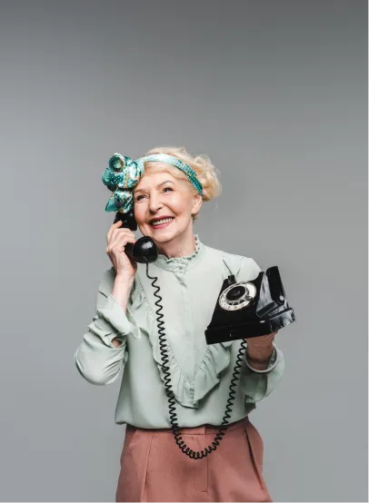 Smiling elderly woman wearing a light green blouse and decorative headband holding a black rotary phone.