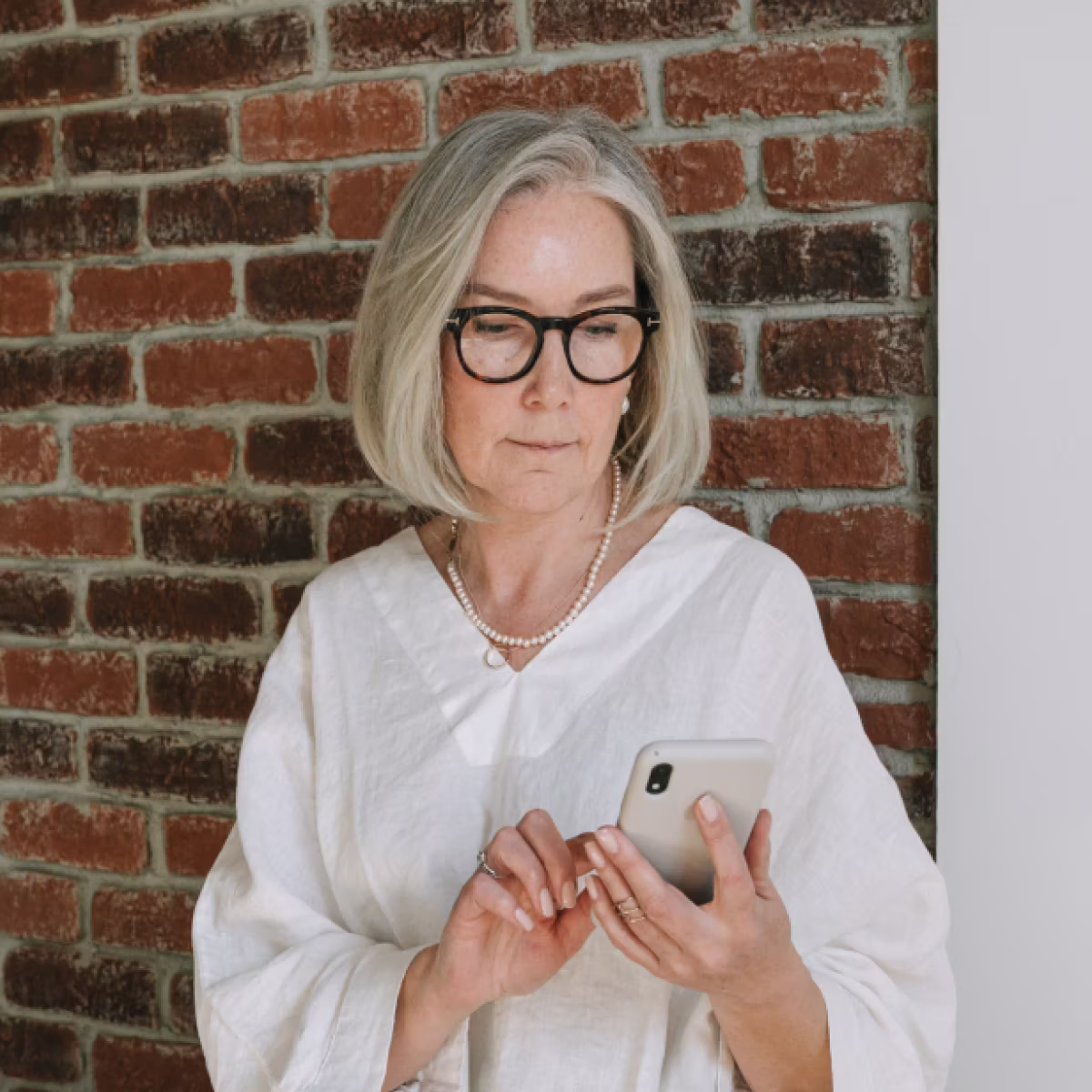 Mature woman with gray hair and glasses in white blouse using smartphone against a brick wall.
