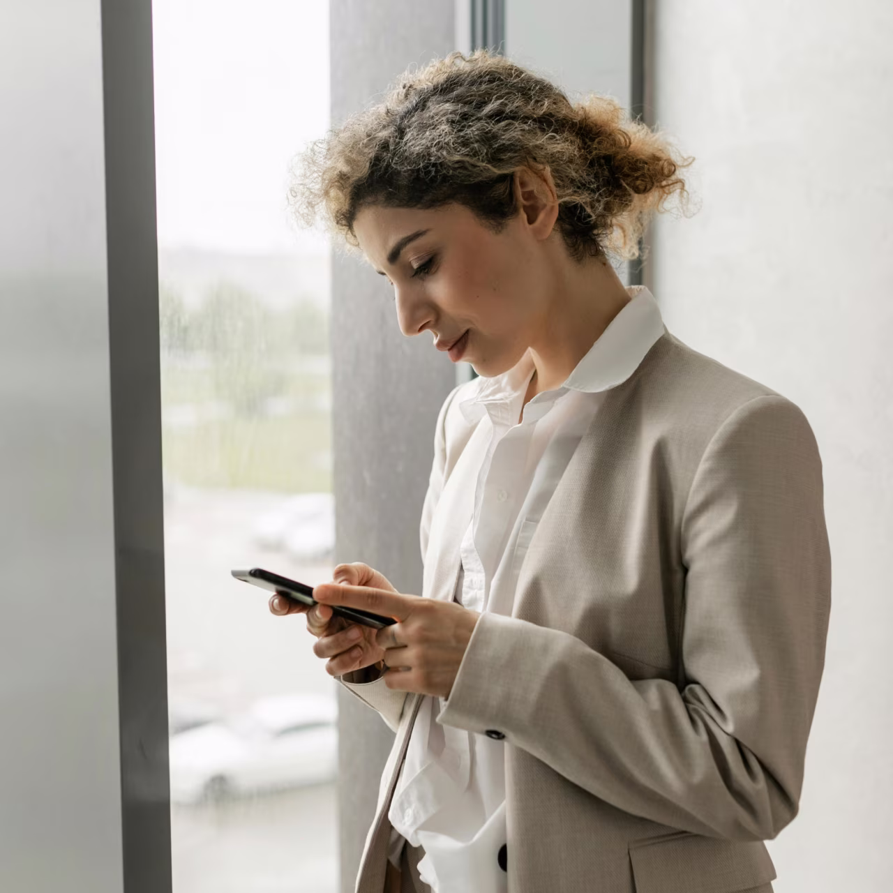 Young woman in beige blazer looking at her smartphone near a window.