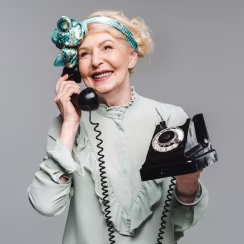 Smiling elderly woman with floral headband holding a black rotary phone handset and base.