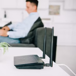 Black Wi-Fi router with two antennas on white table, blurred man sitting on couch in background.