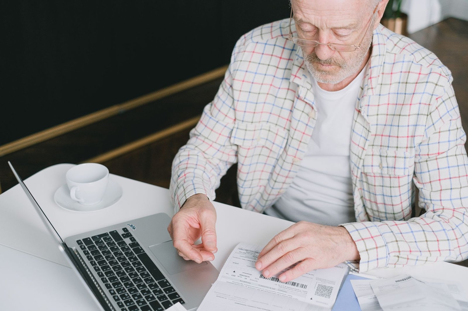 Older man in reading glasses sitting at a table, reading an itemized bill next to a laptop and coffee cup
