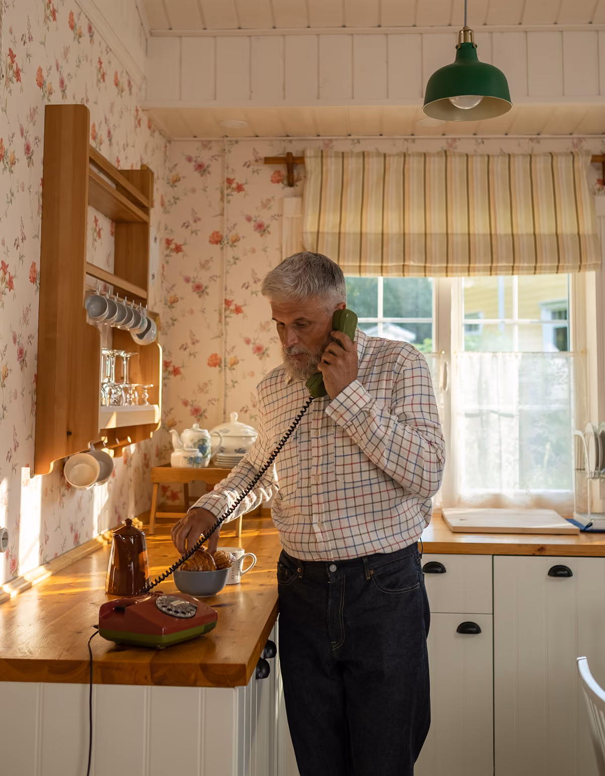 Older man with gray hair and beard standing in a vintage-style kitchen holding a green rotary phone receiver to his ear while resting his other hand on the red rotary phone on the wooden counter, with floral wallpaper, hanging cups, and a window with striped curtains in the background.