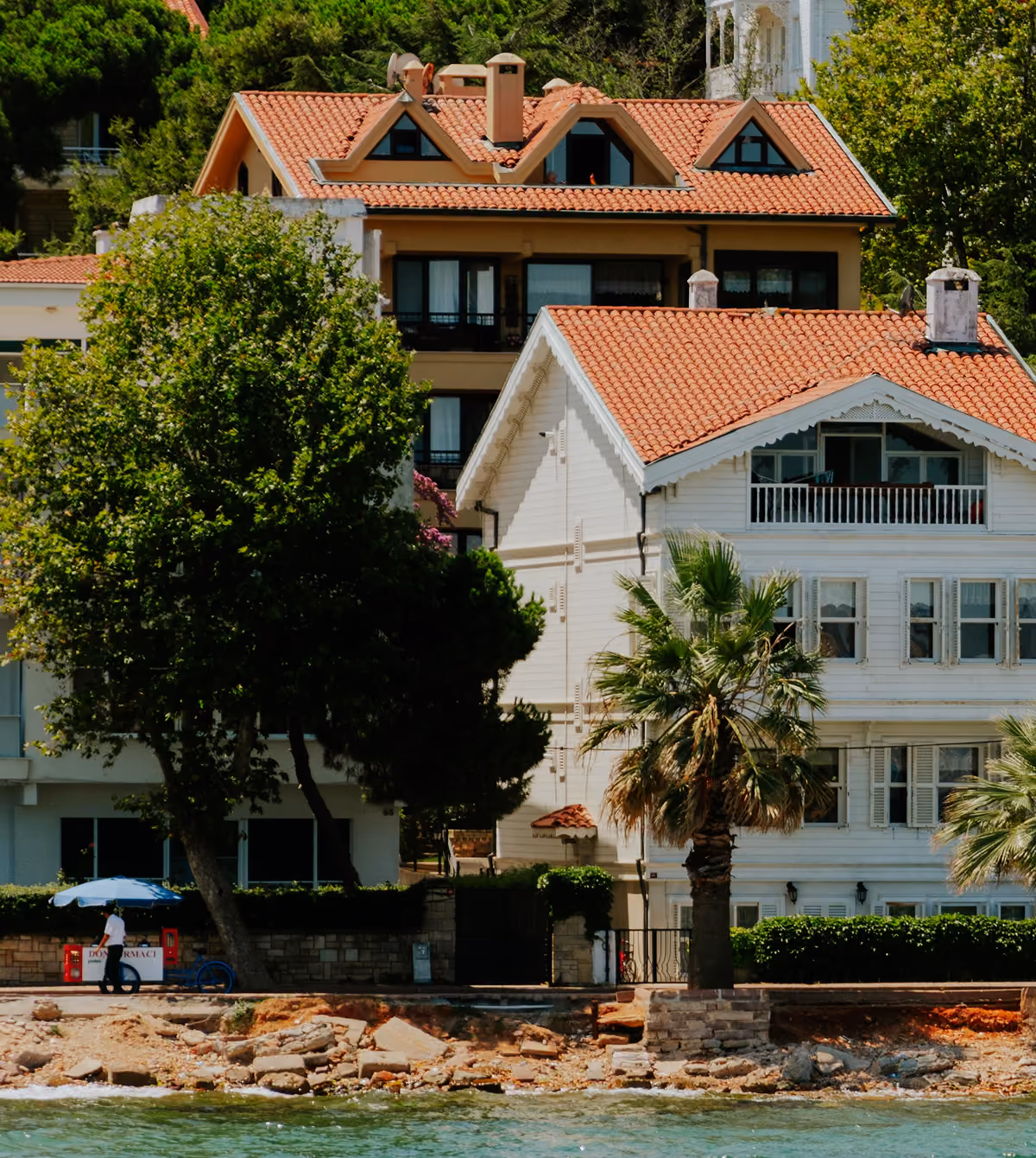 A white two-story house with a red tile roof and decorative trim by the waterfront, surrounded by green trees and a palm tree, with a person walking near a small blue umbrella stand by a rocky shoreline.