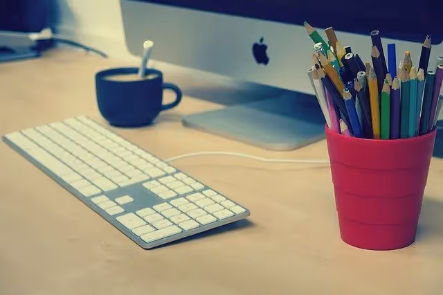 A clean workspace featuring an Apple iMac, a white keyboard, a cup of coffee, and a red container filled with colored pencils.