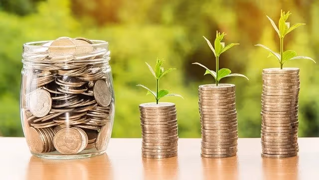 Glass jar filled with coins beside three stacks of coins, each topped with a growing plant.