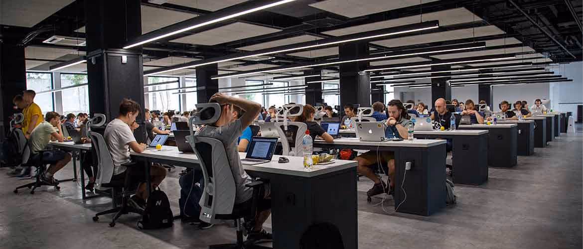 Large open-plan office with many people working at long rows of desks on laptops.