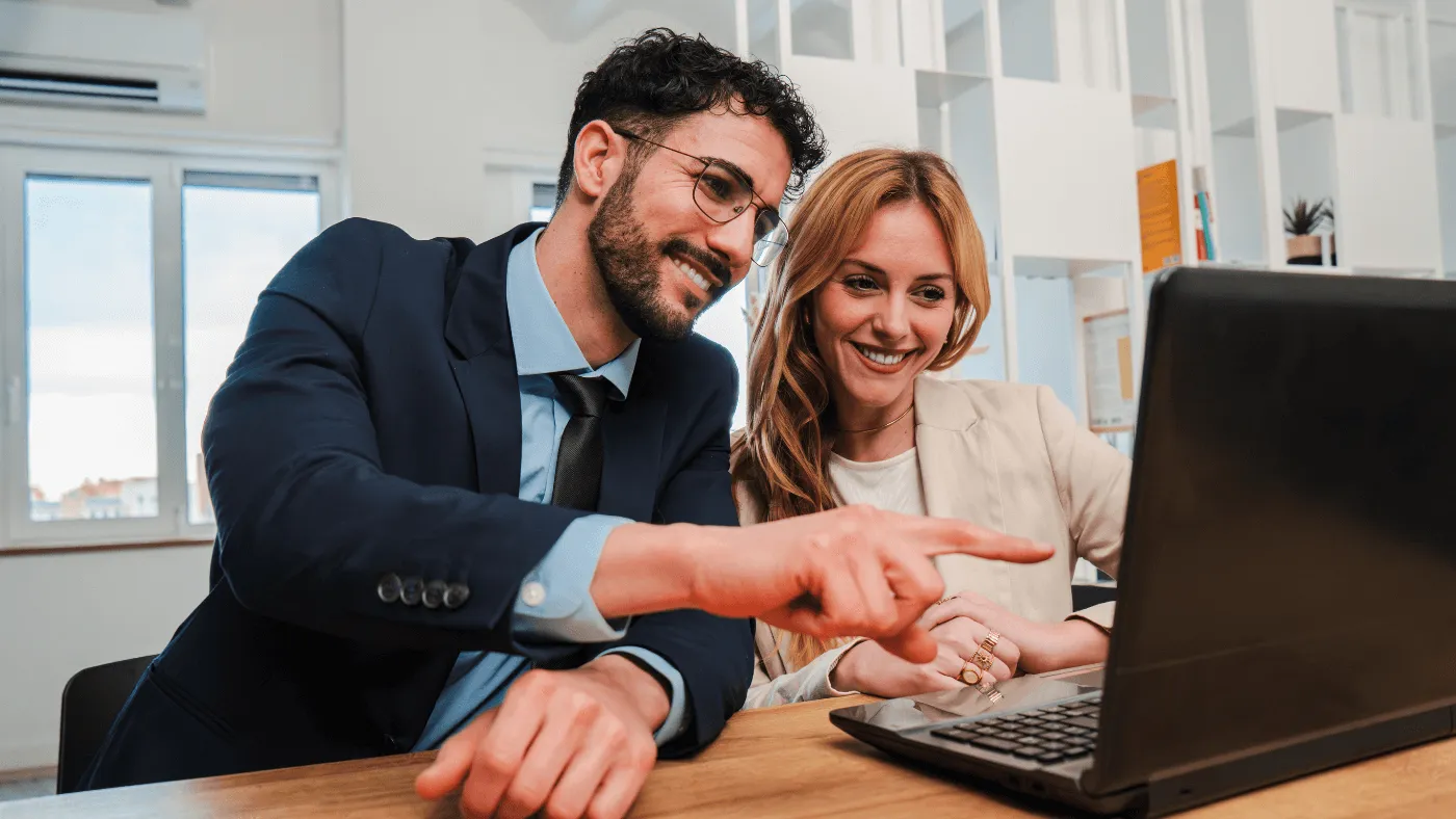A smiling man points at a laptop while sitting next to a smiling woman in an office