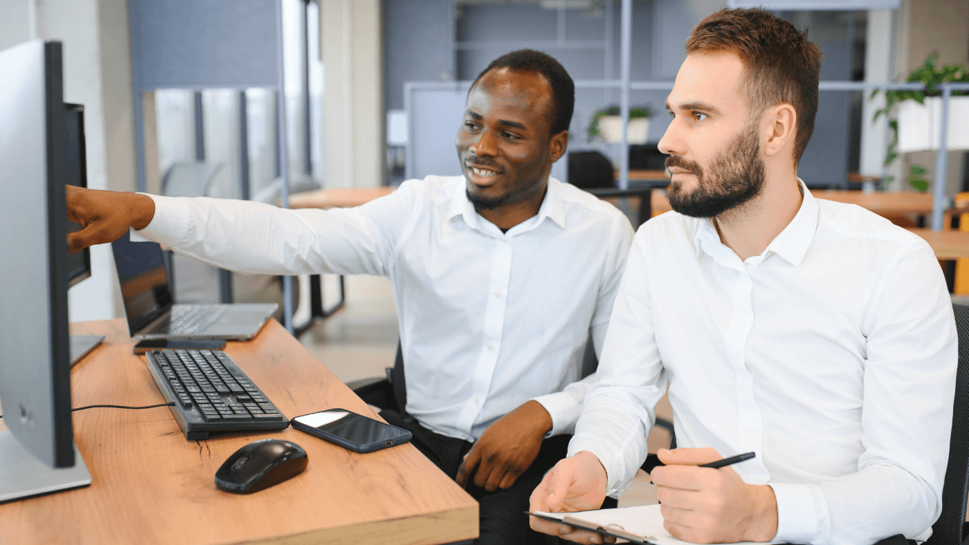 Two men wearing white dress shirts work side-by-side looking at a desktop computer