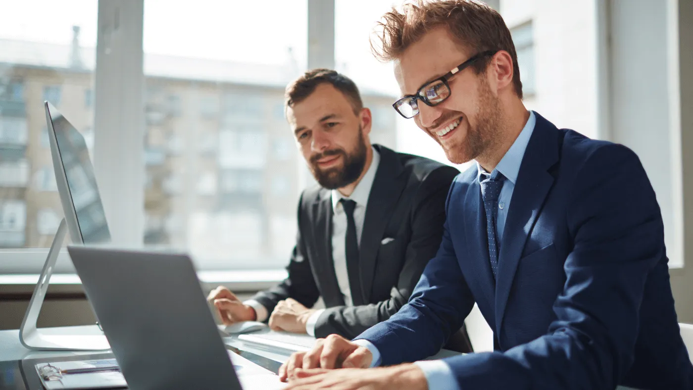 Two men wearing suits and glasses work side by side at a desk on their laptops