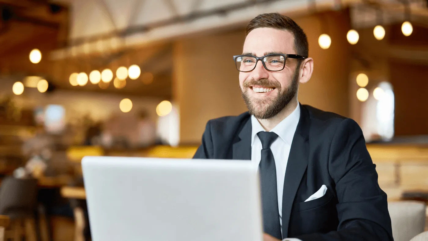 A man wearing a navy suit and tie smiles behind a laptop 