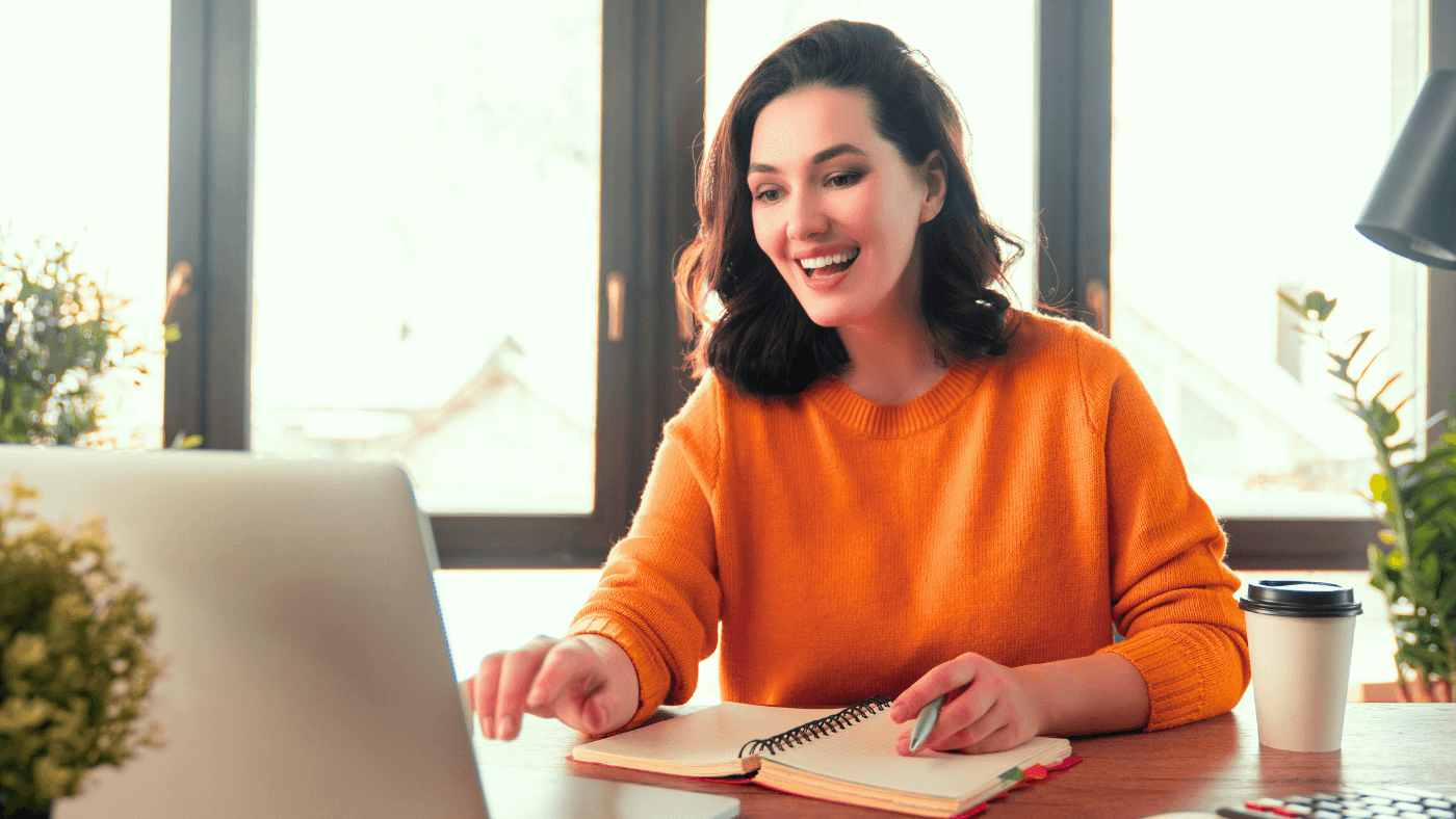 A woman with brown shoulder-length hair wearing a long sleeve orange sweater writes in a notebook in front of an open laptop