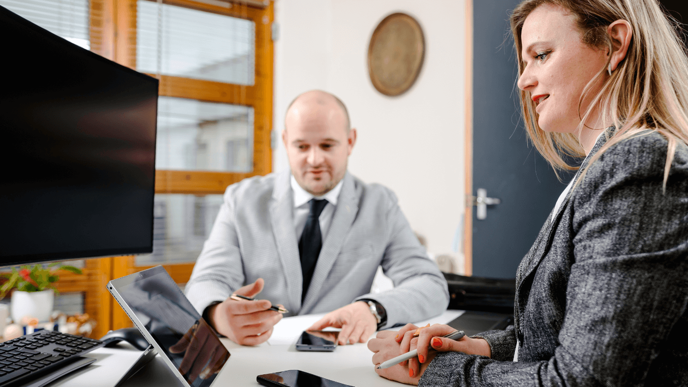A bald man wearing a suit and woman with a blazer work at a desk with their phones and a tablet
