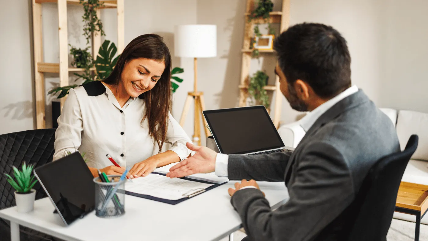 A man wearing a suit sits behind a desk and extends a handshake to a woman wearing a white blazer
