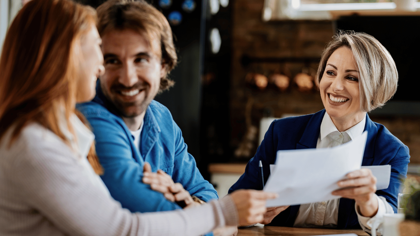 A happy woman wearing a blazer holds papers for a smiling man and woman to review.