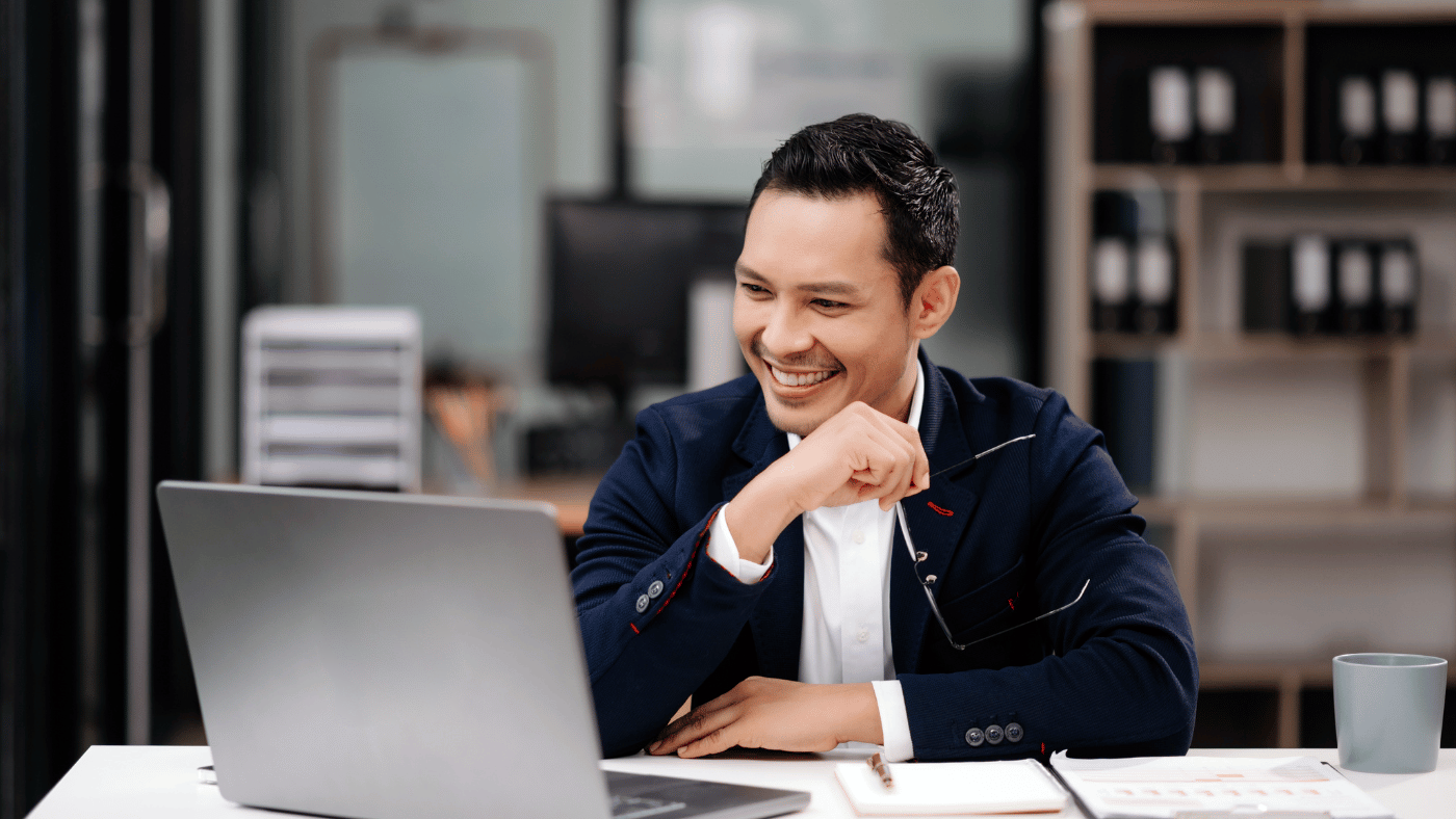 Asian man wearing suit sits at desk and smiles at open laptop.