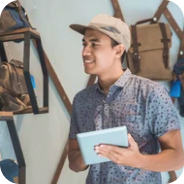 Smiling man in a cap holding a tablet, standing in a store with backpacks displayed on the wall.