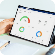 Person holding a tablet displaying dashboard charts including a progress gauge, pie charts, and summary statistics.