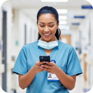 Smiling healthcare worker in blue scrubs looking at a smartphone in a hospital hallway.