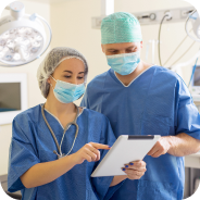 Two healthcare professionals in surgical attire and masks reviewing a tablet in a clinical setting.