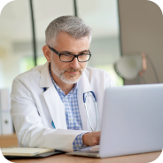 Mature male doctor wearing glasses and a white coat working on a laptop.