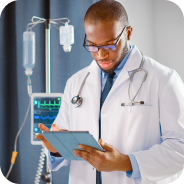 Male doctor wearing white coat and stethoscope, reviewing a tablet in a hospital room with medical equipment in the background.