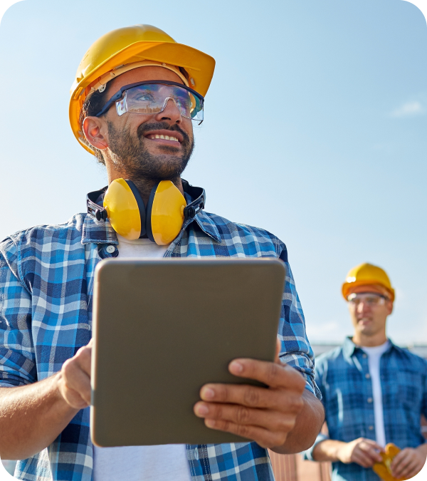 Construction worker wearing a yellow hard hat, safety glasses, and ear protection, holding a tablet with another worker in the background.