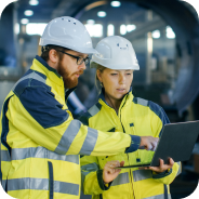 Two industrial workers in yellow safety jackets and white helmets reviewing information on a laptop in a factory setting.