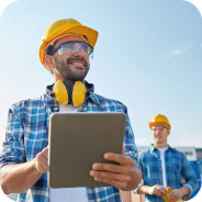 Smiling construction worker wearing a yellow helmet holding a tablet with another worker in the background.