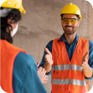 Two construction workers wearing yellow helmets and orange safety vests discussing at a worksite.