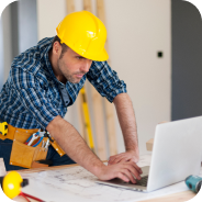 Construction worker in a yellow hard hat and plaid shirt working on a laptop with building plans on a table.