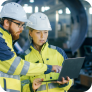 Two engineers in white helmets and yellow safety jackets reviewing a laptop in an industrial setting.