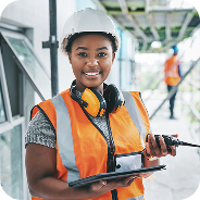 Smiling construction worker wearing a white hard hat, orange safety vest, and yellow headphones holding a tablet at a building site.