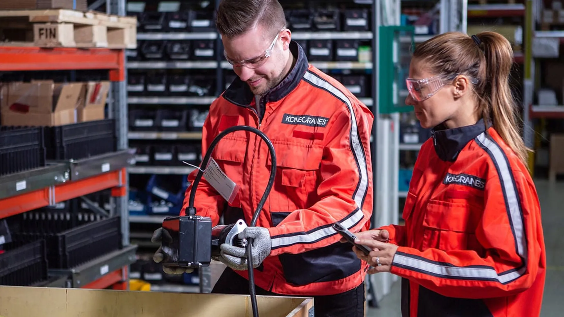 Two Konecranes technicians in orange work jackets and safety glasses inspecting a spare part in a parts warehouse, one checking details on a phone