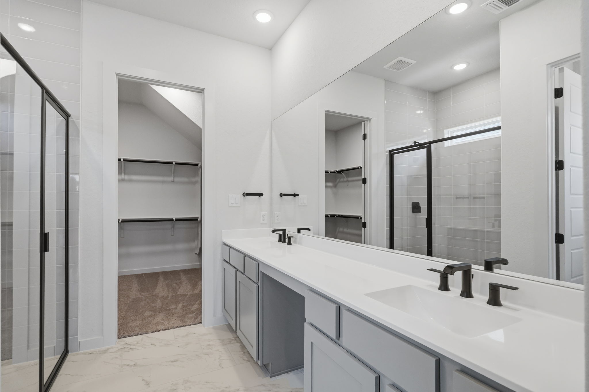 Primary suite bathroom in The Travis floorplan featuring a long double vanity with dark matte black fixtures, full-length mirror, marble-look tile flooring, framed glass shower, and entrance to a walk-in closet.