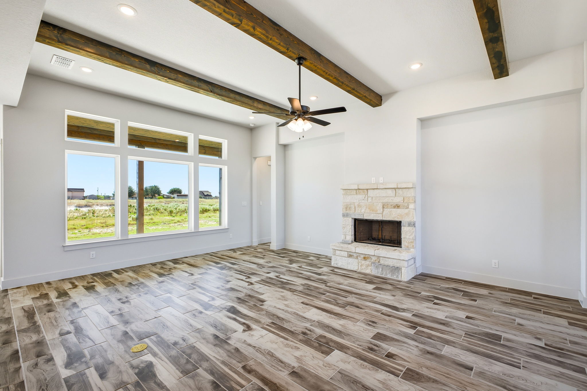 Open-concept living room featuring exposed dark timber ceiling beams, a floor-to-ceiling stone fireplace, and wide-plank wood-look flooring.