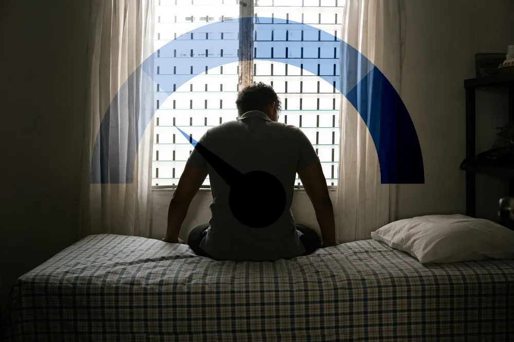 Mann mit Hypogonadismus sitzt niedergeschlagen auf dem Bett, vor dem Fenster erscheint eine leere Skala als Symbol für Testosteronmangel, Erschöpfung und Antriebslosigkeit. | GoSpring