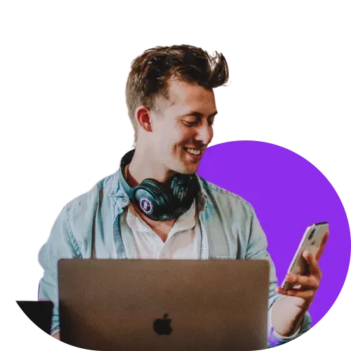 Man with headphones around his neck, smiling at his smartphone while working on a laptop