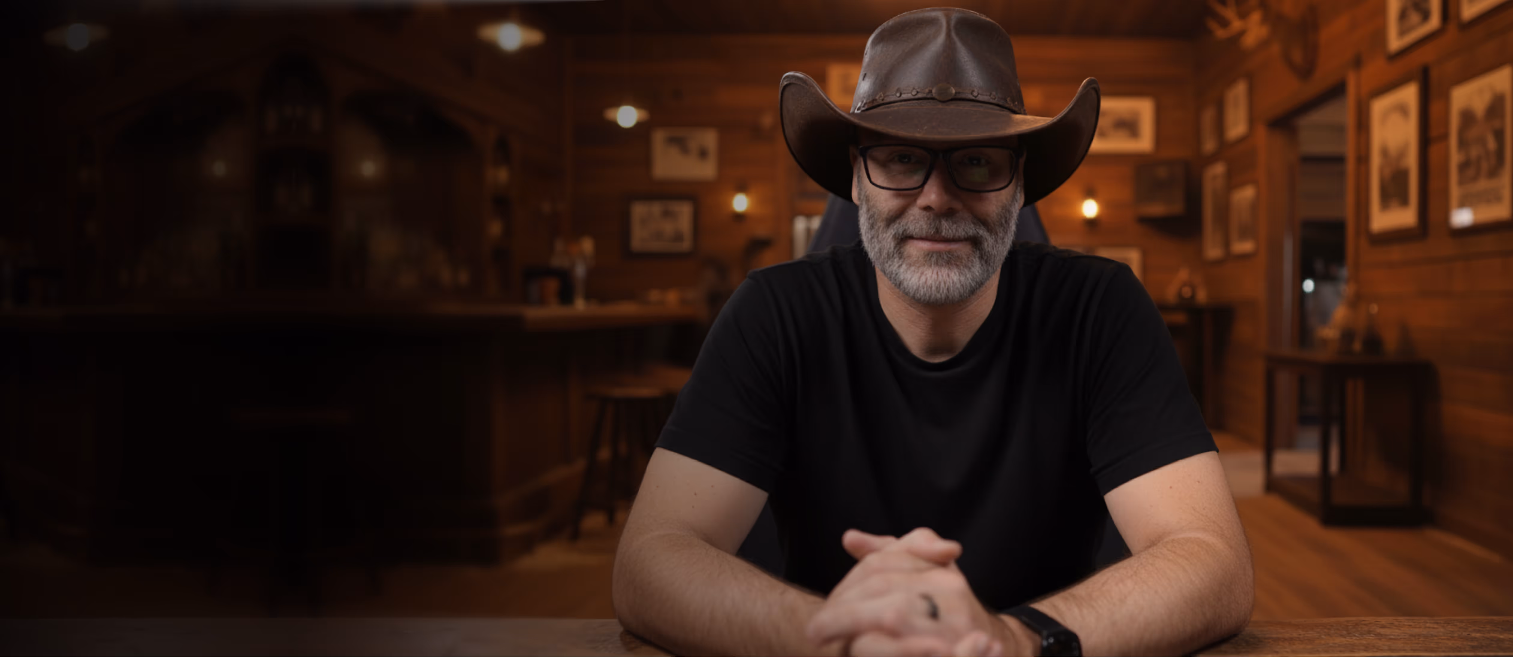 Man wearing a cowboy hat and glasses sitting at a wooden bar interior, looking at the camera