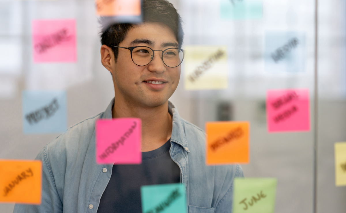A man wearing glasses and a denim shirt looks at colorful sticky notes on a glass wall during a planning or brainstorming session.