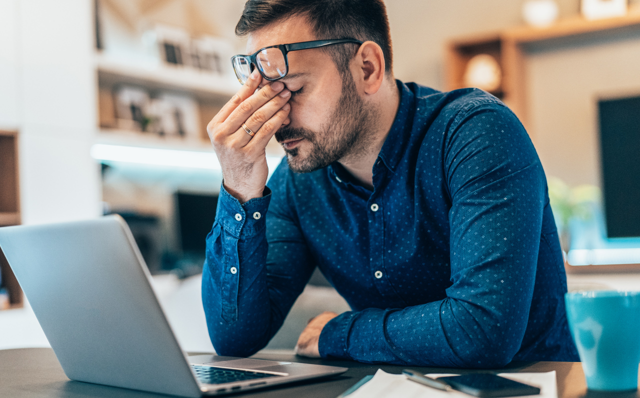 Man sitting at a desk with a laptop, wearing glasses and a blue shirt, holding his face in frustration or exhaustion.