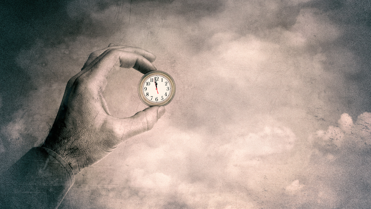 A close-up of a person’s hand holding a small round clock against a cloudy sky background, symbolising the passing of time or a countdown. The muted, vintage tones convey urgency and decay, aligning with the idea that time is running out.