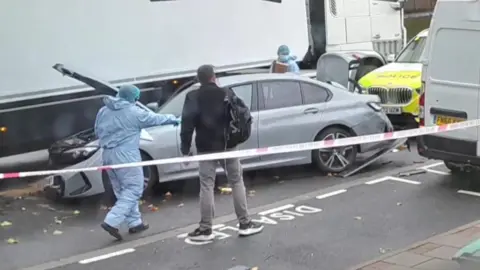 A silver car is pictured crashed into a lorry. Its boot is up and a police car is parked immediately behind it. Police tape is around the scene and a forensic officer wearing a blue hazmat suit is carrying equipment over their shoulder while pointing to the car. Another forensics officer is at the back of the car clutching a box.
