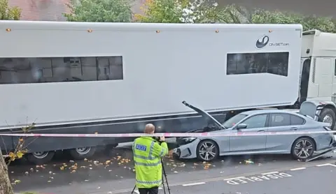 A policeman takes a photo of a grey car that has crashed and is wedged, with it's boot lid forced open, under a grey truck. In front is a red police tape.