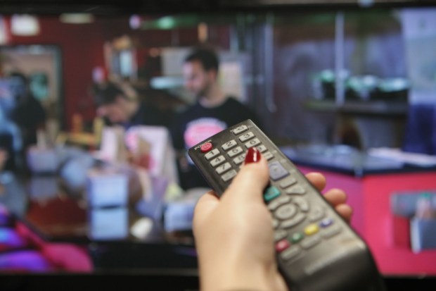 A hand holding a remote control pointing at a TV screen showing a man in a black t-shirt.